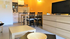 A modern kitchen with white cabinets, a dining area with a wooden table and chairs, and a flat-screen TV on a wooden stand. - New apartment 2/4 people, 100m to beach, Perros-Guirec (Perros-Guirec)