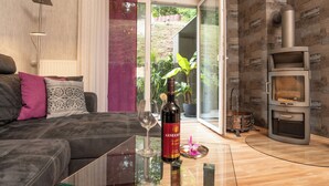 Ein modernes Wohnzimmer mit einer Couch, einer Weinflasche und einem Glas. Eine Glastür öffnet sich zu einem Außenbereich mit Pflanzen.