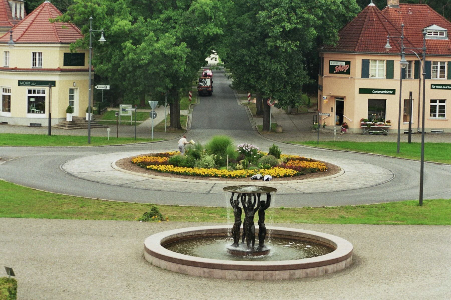 Ein Kreisverkehr mit einer zentralen Fontäne und Blumenbeeten, umgeben von Gebäuden mit Beschilderung.