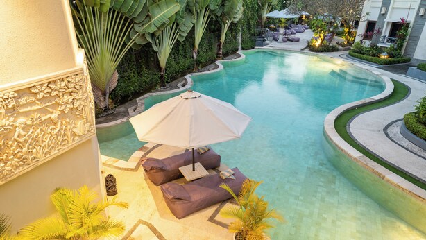 Una piscina curva con una zona de estar que incluye una sombrilla color beige y cojines.
