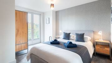 A modern hotel room with a large bed, a wooden wardrobe, a grey headboard, and a nightstand with a lamp.