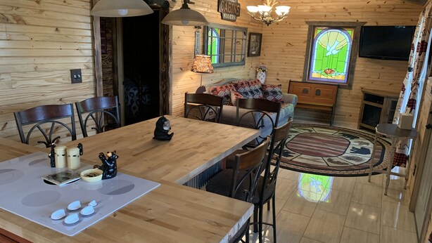 Interior de uma cabana de madeira com uma grande mesa de jantar, cadeiras, cozinha e uma área de estar acolhedora com um tapete.