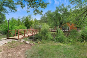 Un pont en bois enjambe un ruisseau et mène à un chalet rustique entouré d’une forêt dense.
