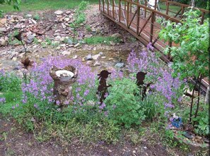 Cross a tranquil wooden bridge over a serene garden stream, surrounded by lush greenery and vibrant flowers.