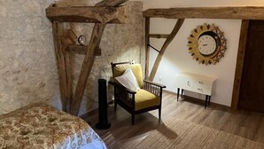 A rustic room with exposed wooden beams, a bed with patterned bedding, a yellow armchair, a white bedside table, and a wall clock. - Luxury apartment in a renovated Chateau with a  view of the Pyrenees. (Mouzens)