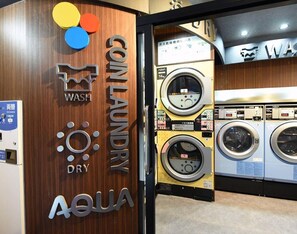 Une laverie équipée de plusieurs machines à laver et de plusieurs séchoirs, un mur en bois sur lequel on peut lire les inscriptions « Laverie automatique » et le nom de la marque « Aqua », ainsi qu’un panneau indiquant la présence d’un service d’échange.