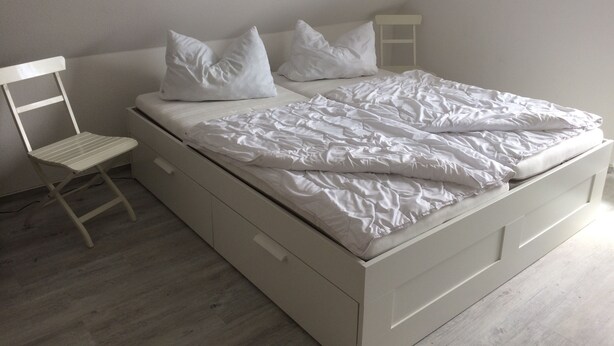 Een wit bed met kussens en een witte stoel in een kamer met een schuine plafond.