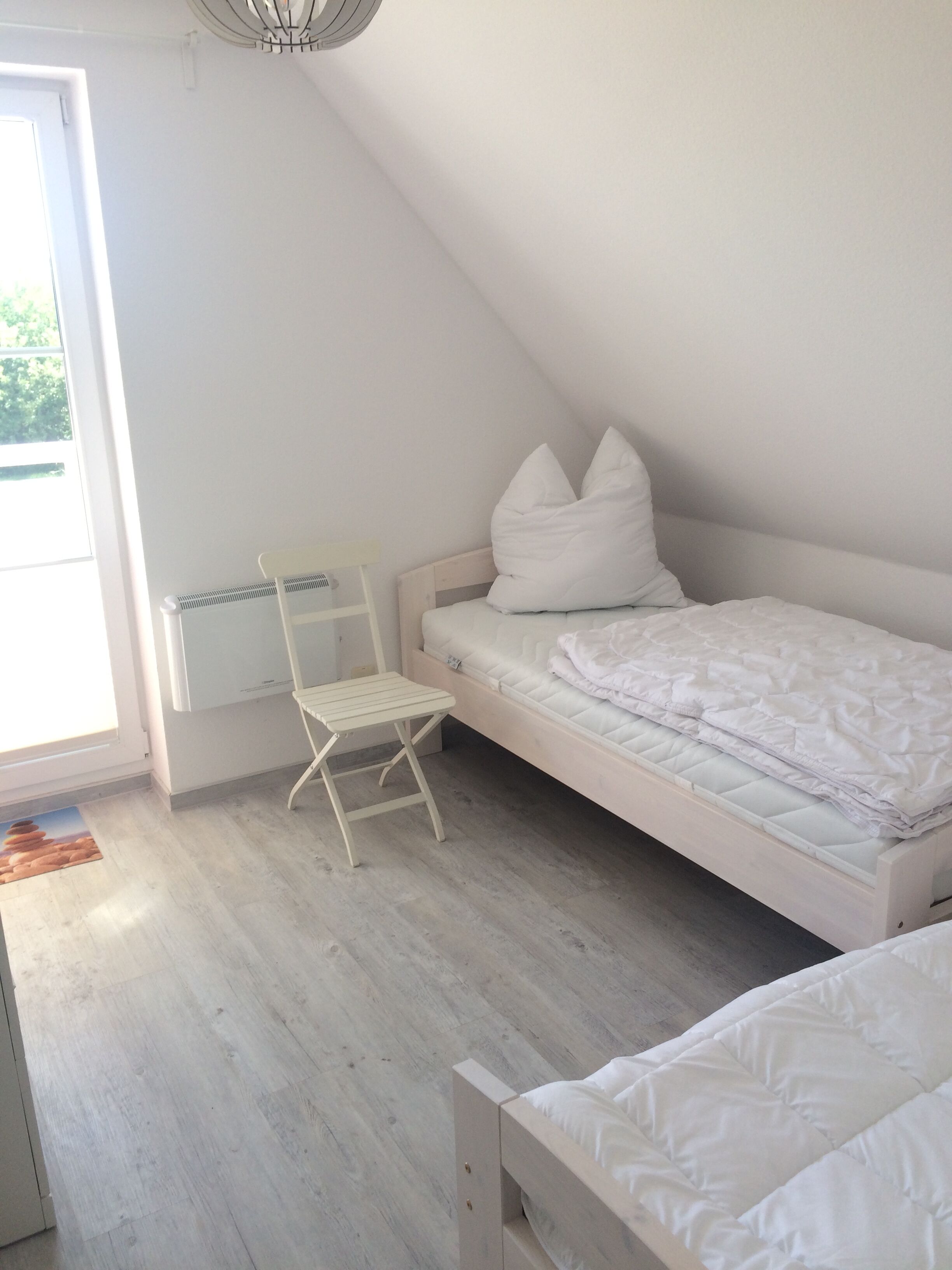 A bedroom with a sloped ceiling, a single bed with white bedding, a small white chair, and a radiator.