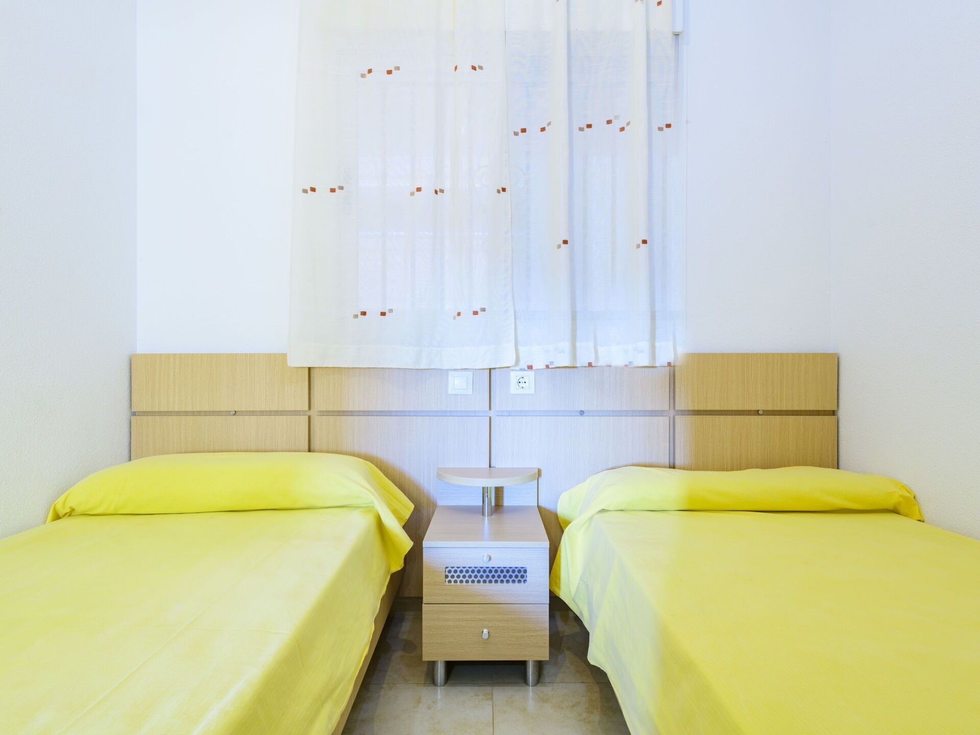 A room with two beds, each with a yellow bedspread, a wooden headboard, and a small bedside table with a drawer.