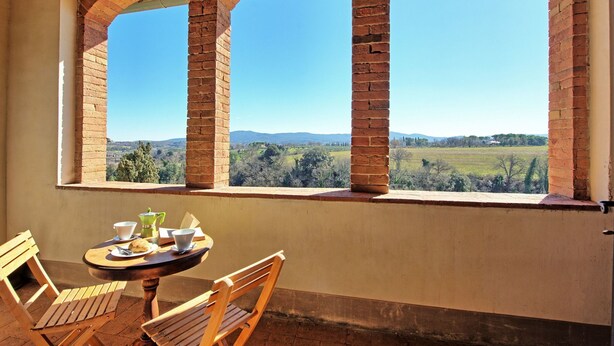 Quarto com uma mesa, cadeiras, janela com vista para um campo e colinas, e uma parede de tijolos.