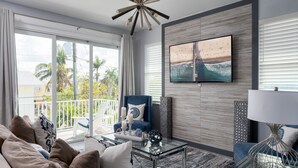 A modern living room with a large TV, comfortable seating, and a balcony with a view of palm trees. - Modern 4BR Beach House | Pool, Walk to Beach & Prime Location (Fort Myers Beach)