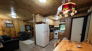 A cozy cabin interior with wooden walls, a dining area with a table and chairs, a kitchen with a refrigerator and stove, and a living room with a sofa and a lamp. - Cardinal's Landing-Covered Porch - Pet-Friendly - Private-4WD-AWD (Bryson City)