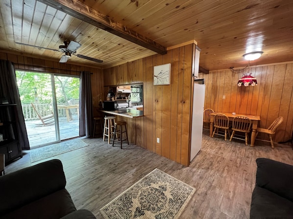 A cozy cabin interior with a wooden ceiling, a dining area with four chairs, a kitchen with a refrigerator, and a large window overlooking a deck.