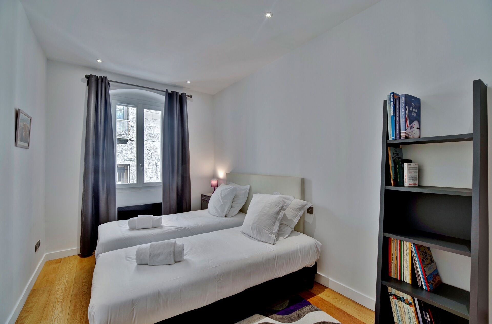 A hotel room with two single beds, a bookshelf with books, a window with curtains, and a wooden floor.