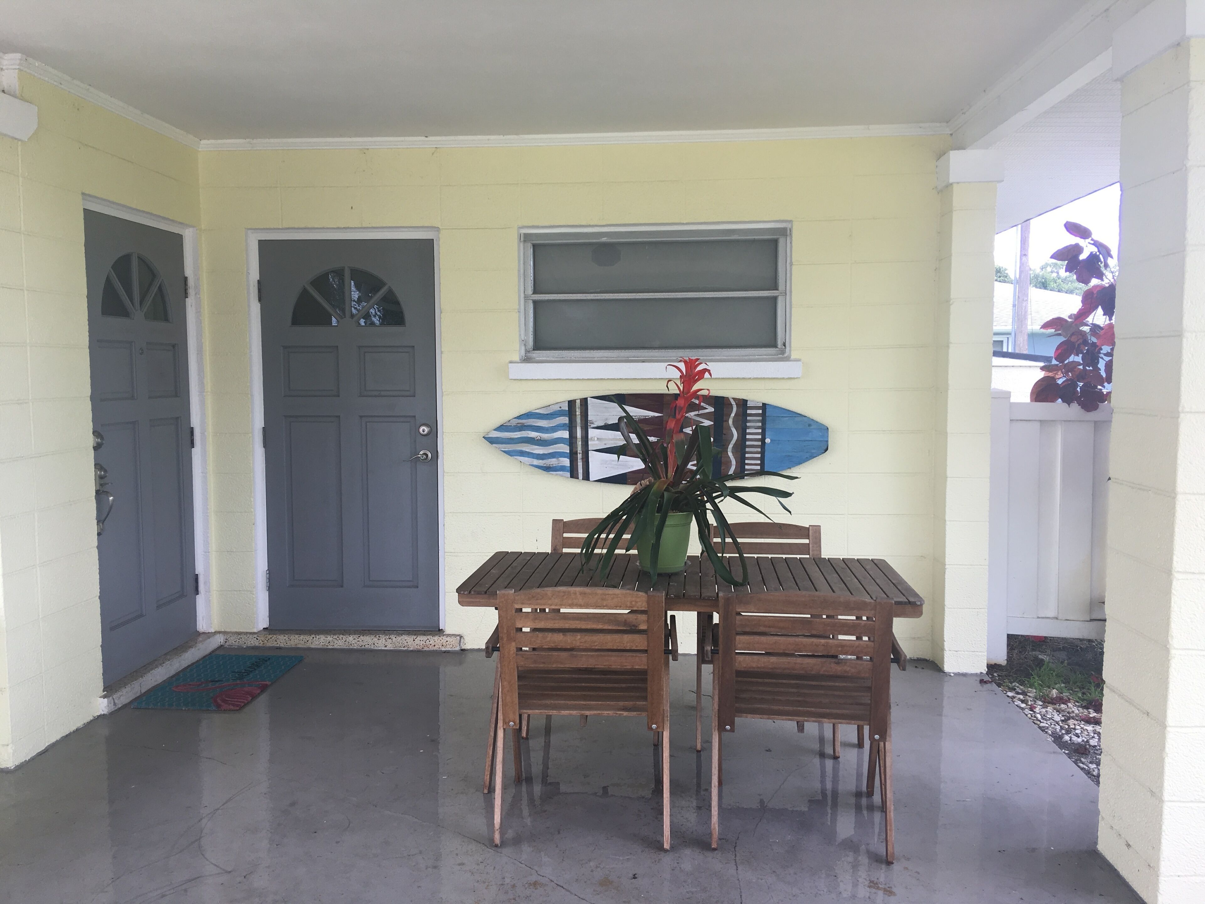 A porch with a wooden table and chairs, a potted plant, a surfboard decoration, and a door.