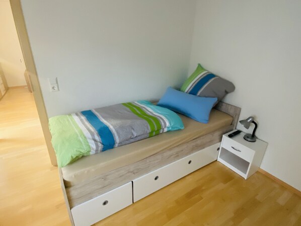 A small, single-bed room with a wooden floor, a white bedside table with a lamp, and a striped blanket.