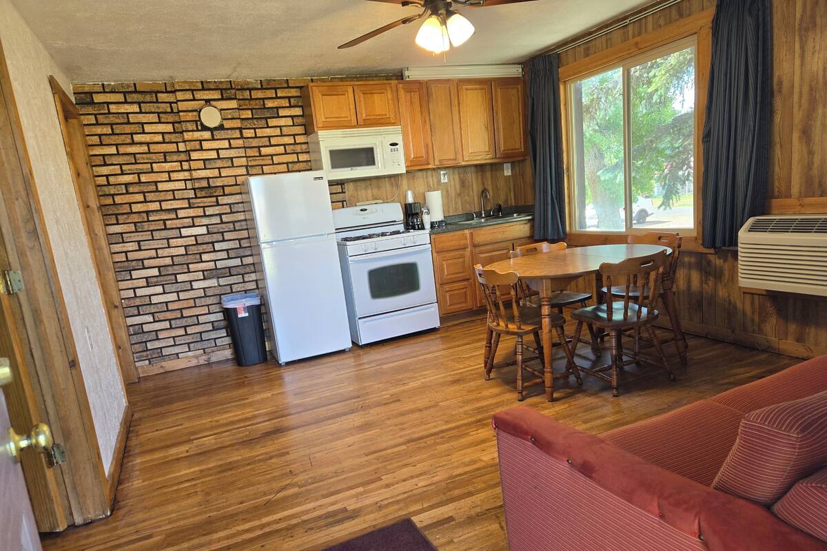 A compact kitchen with a refrigerator, stove, microwave, and wooden cabinets. There's a dining table with chairs and a red couch in the living area.