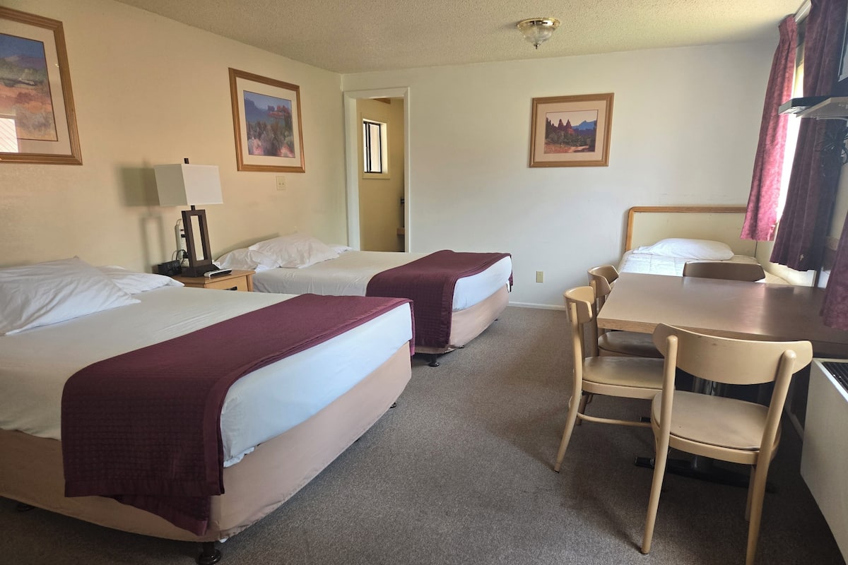 A hotel room with two beds, a desk, and a chair. There are framed pictures on the walls and a window with curtains.