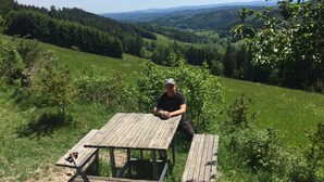 Ein Mann sitzt an einem Holztisch in einem grasbewachsen Feld mit Blick auf Hügel und Bäume im Hintergrund.