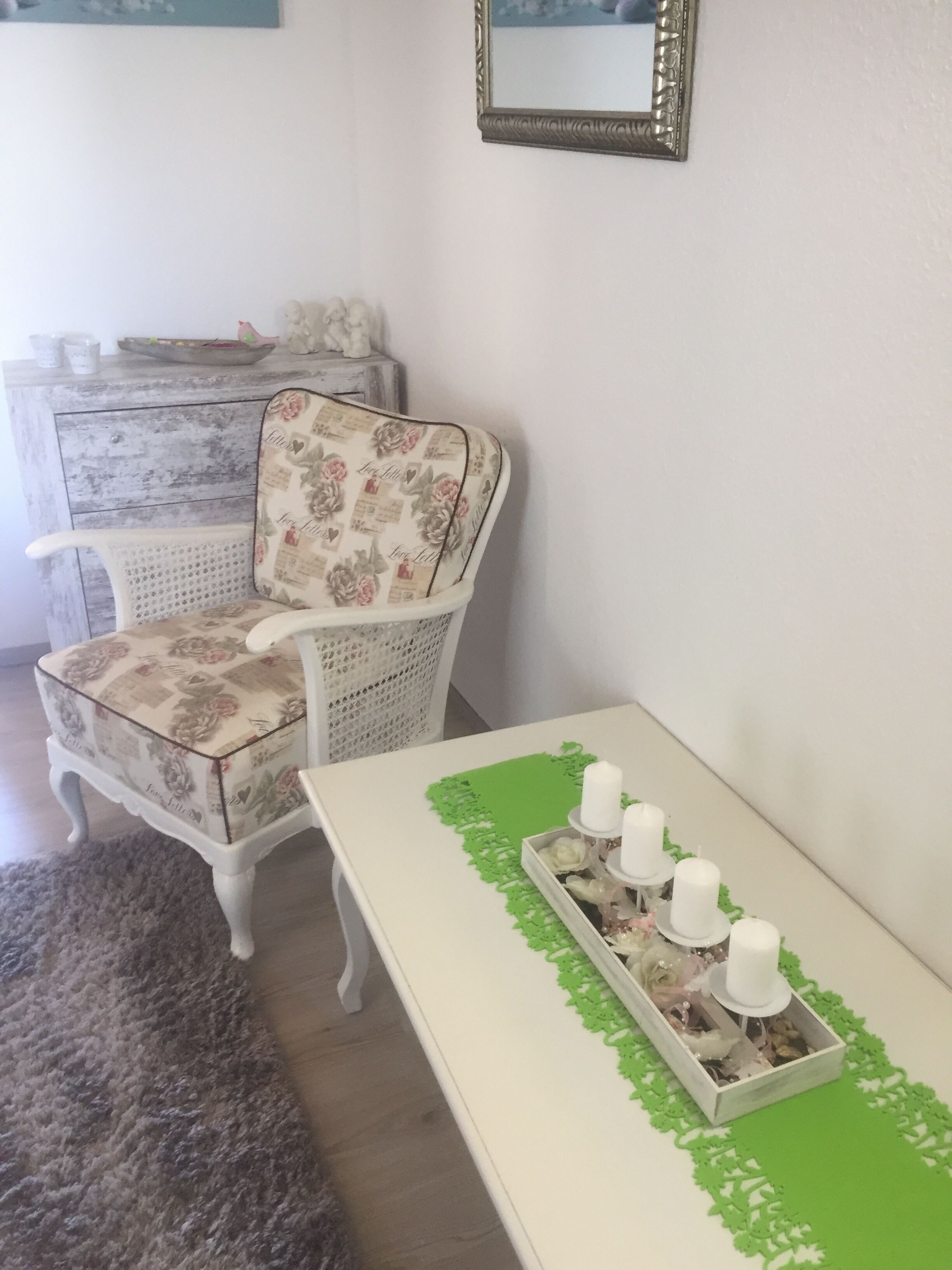 A room with a white chair with floral upholstery, a white table with a green doily, and a mirror on the wall.