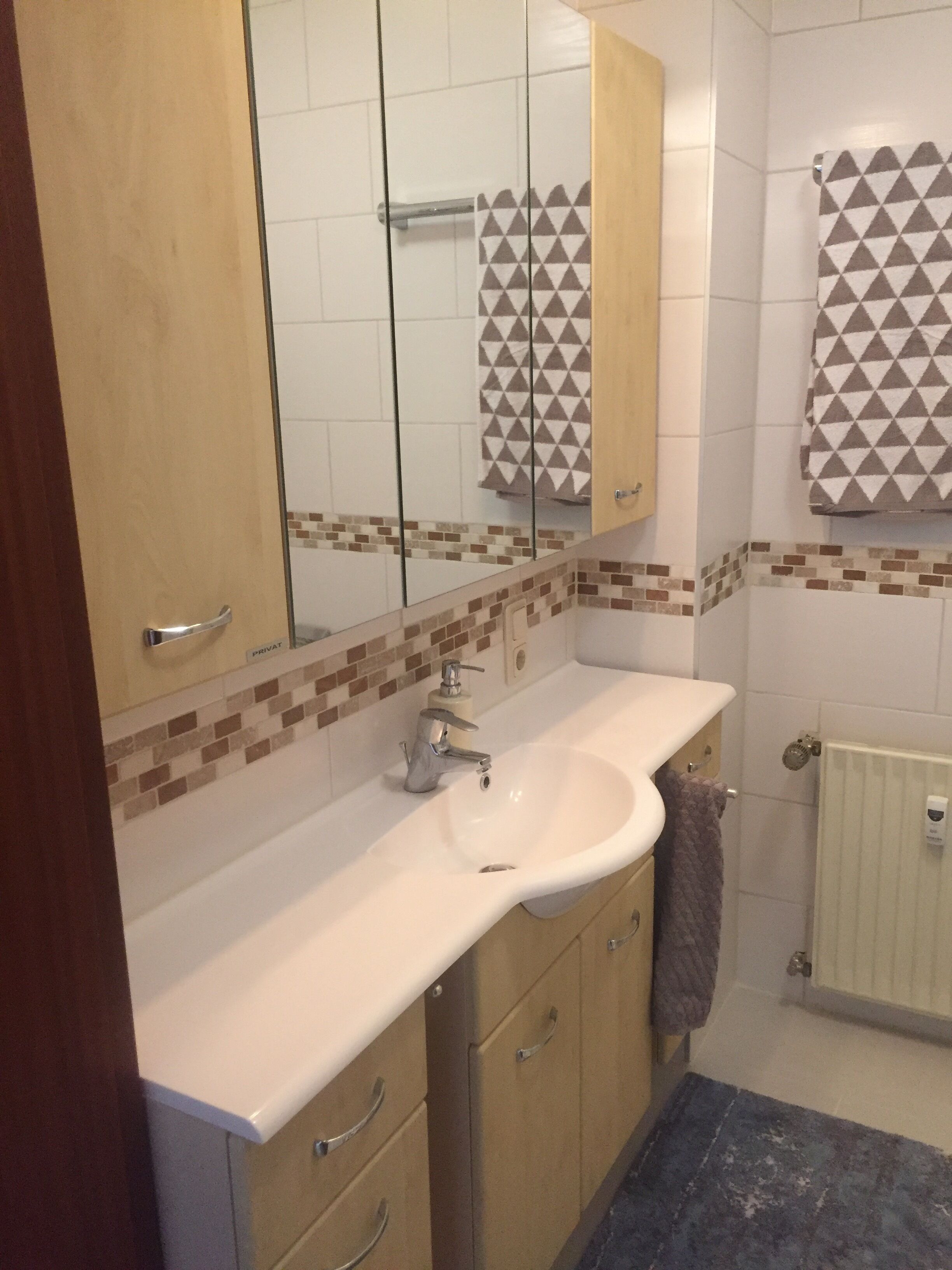 Une salle de bain avec un lavabo blanc, un miroir, un porte-serviettes et un mur carrelé orné d’un motif géométrique.