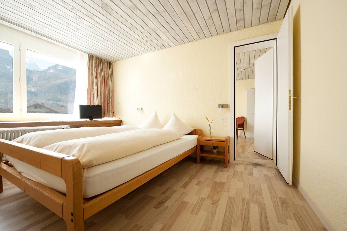 A bedroom with a wooden bed, a bedside table, a lamp, and a view of mountains through a window.