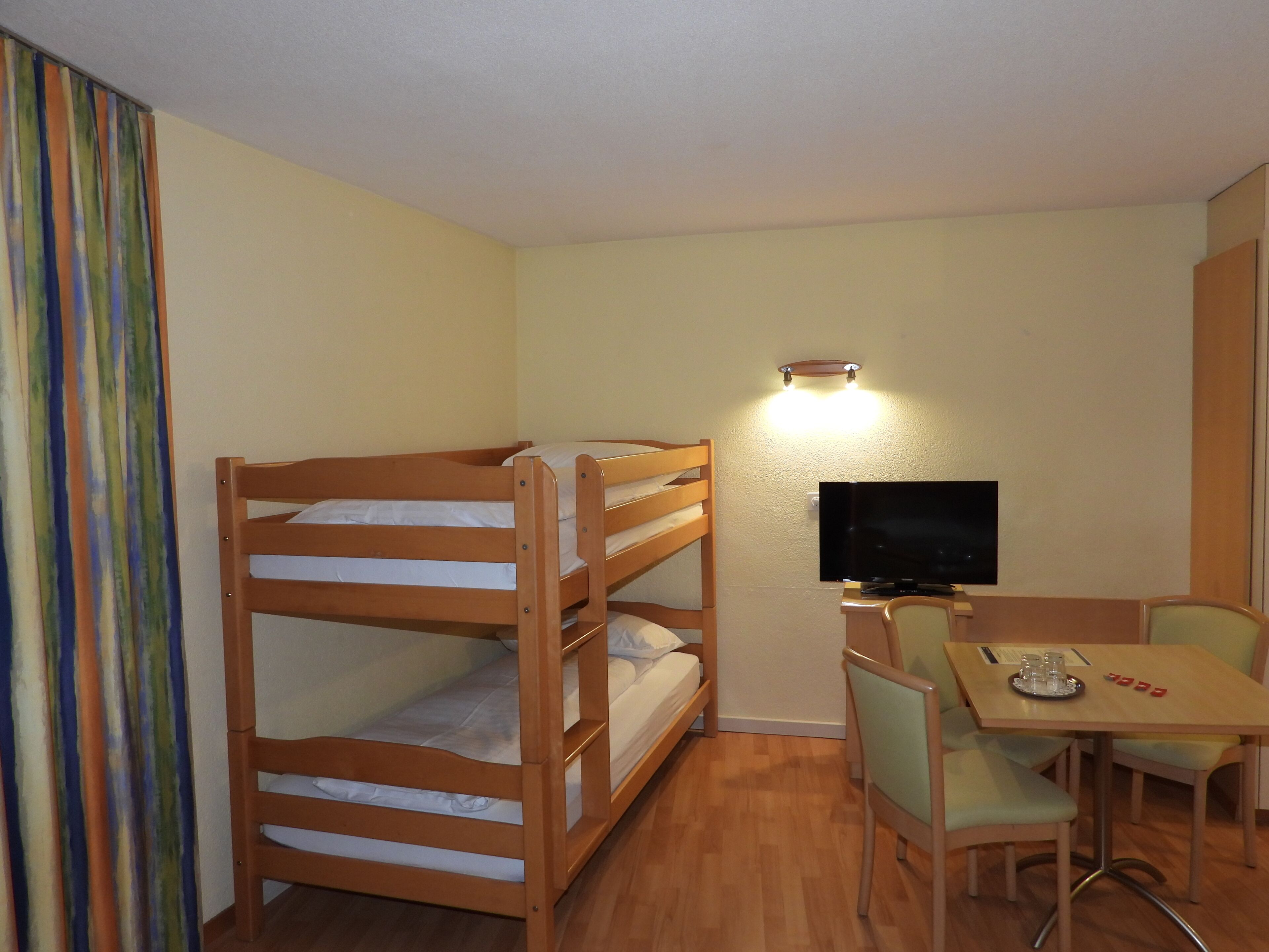 A room with bunk beds, a desk with a chair, a television, and a curtain.