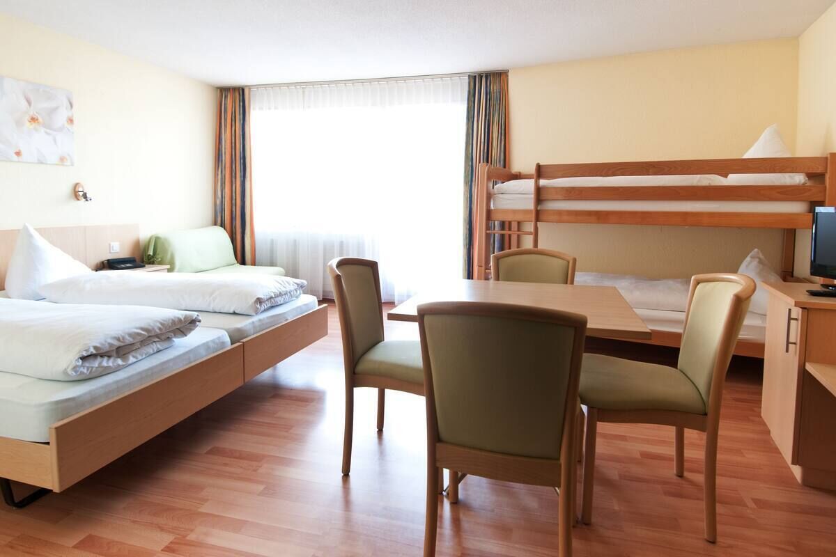 A hotel room with a bunk bed, a desk with a chair, and a bed with a bedside table.