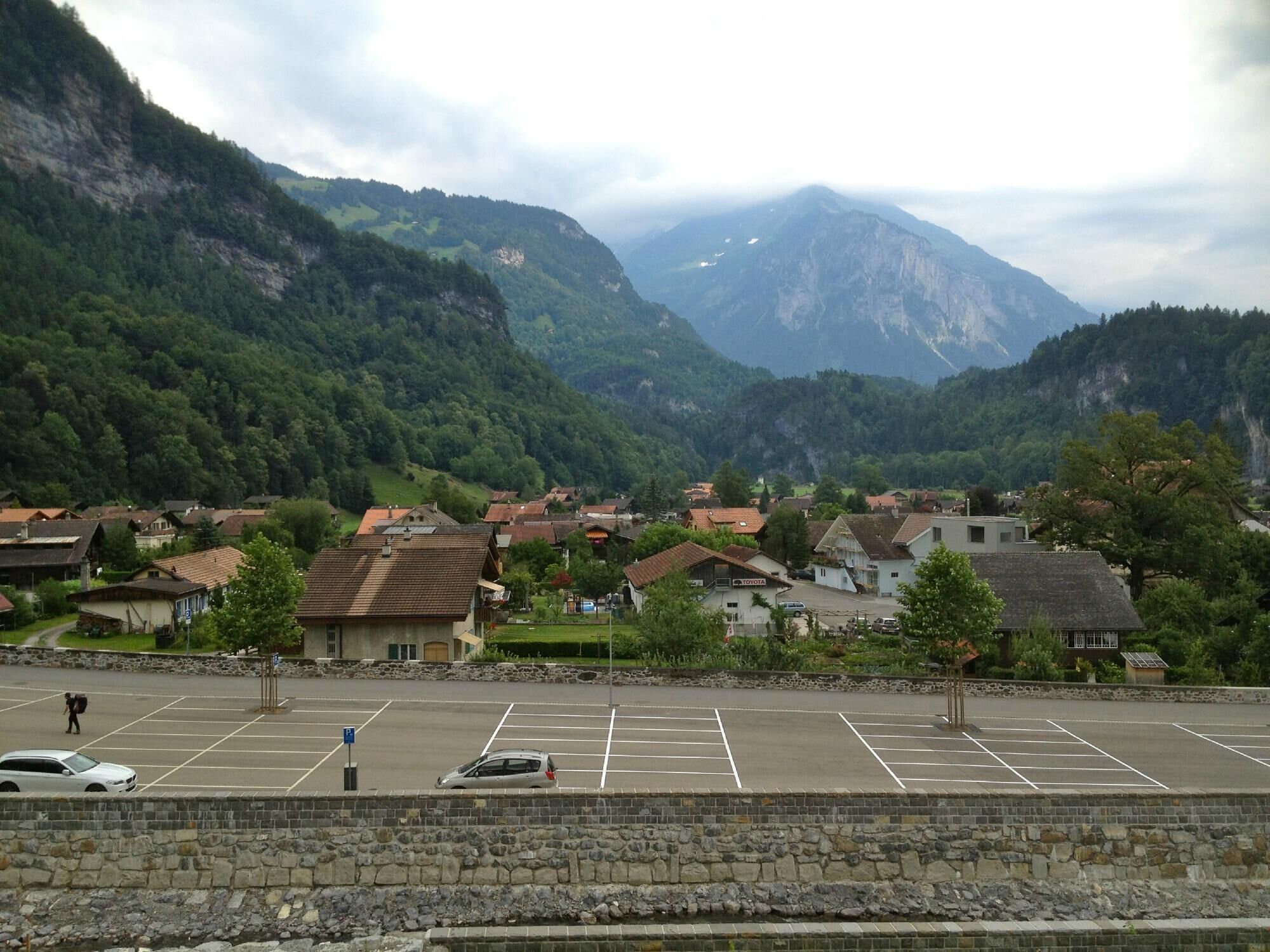 A parking lot with cars, a stone wall, and a view of a village with houses and mountains in the background.
