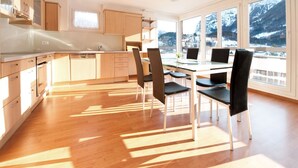 A modern kitchen with wooden cabinets, a dining area with black chairs, and large windows offering a view of snow-capped mountains. - Hotel Sherlock Holmes "nur 200 m zur Gondelbahn Hasliberg" (Meiringen)
