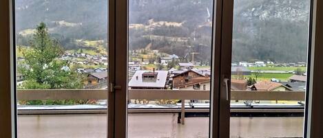 A view through a window with a balcony, overlooking a mountainous landscape with houses and a small town.
