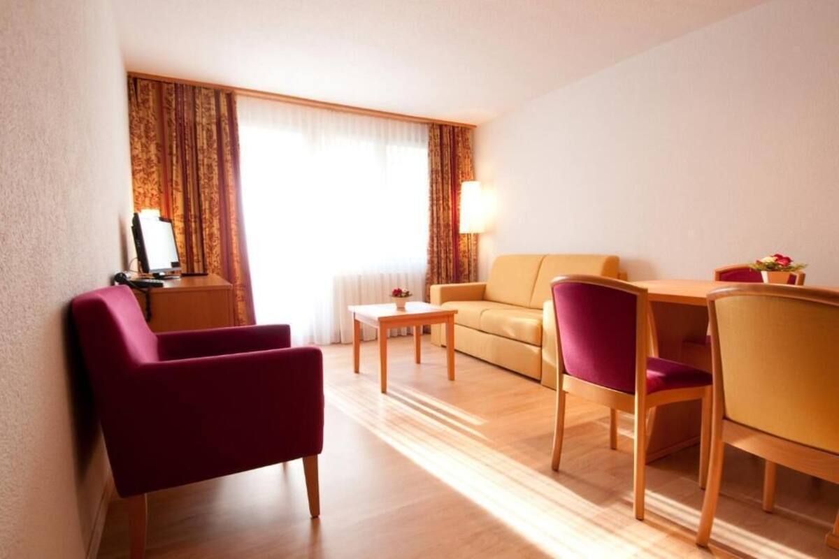 A hotel room with a sofa, two armchairs, a dining table with chairs, a lamp, and a window with curtains.