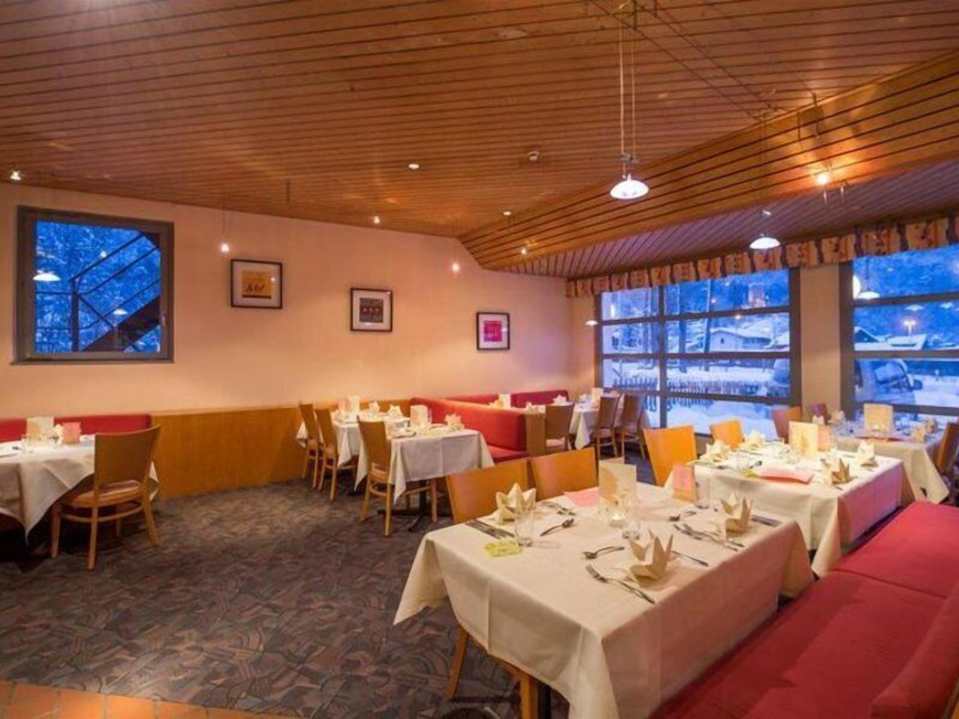 Un restaurante con techo de madera, mesas dispuestas para comer y grandes ventanales con vistas a un paisaje nevado.