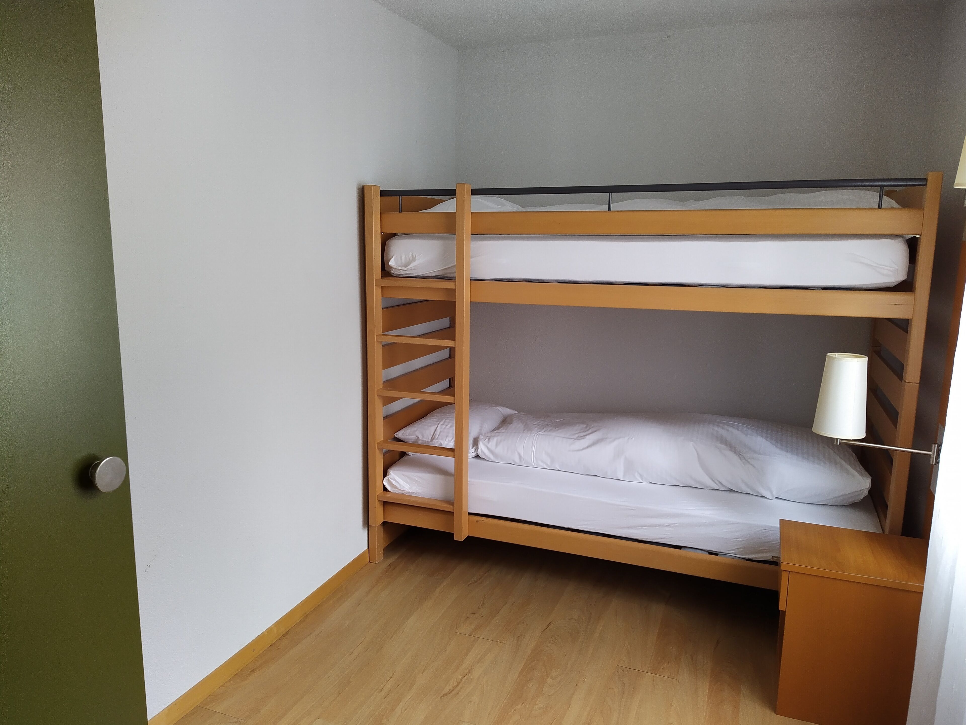 A bunk bed with a wooden frame and white bedding in a corner room with a wooden floor and a green wall.