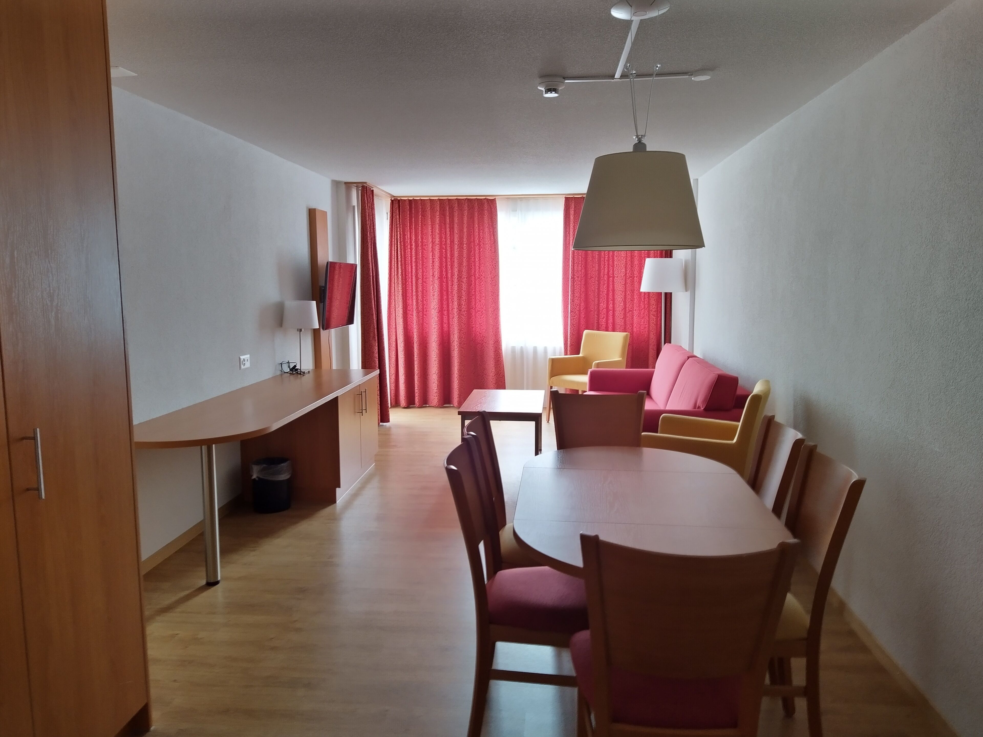 Een moderne, goed verlichte kamer met een eettafel, stoelen, een bank en een aan de muur bevestigde televisie.