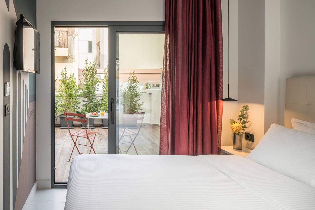 A hotel room with a bed, a desk, a chair, and a view of the outside balcony.