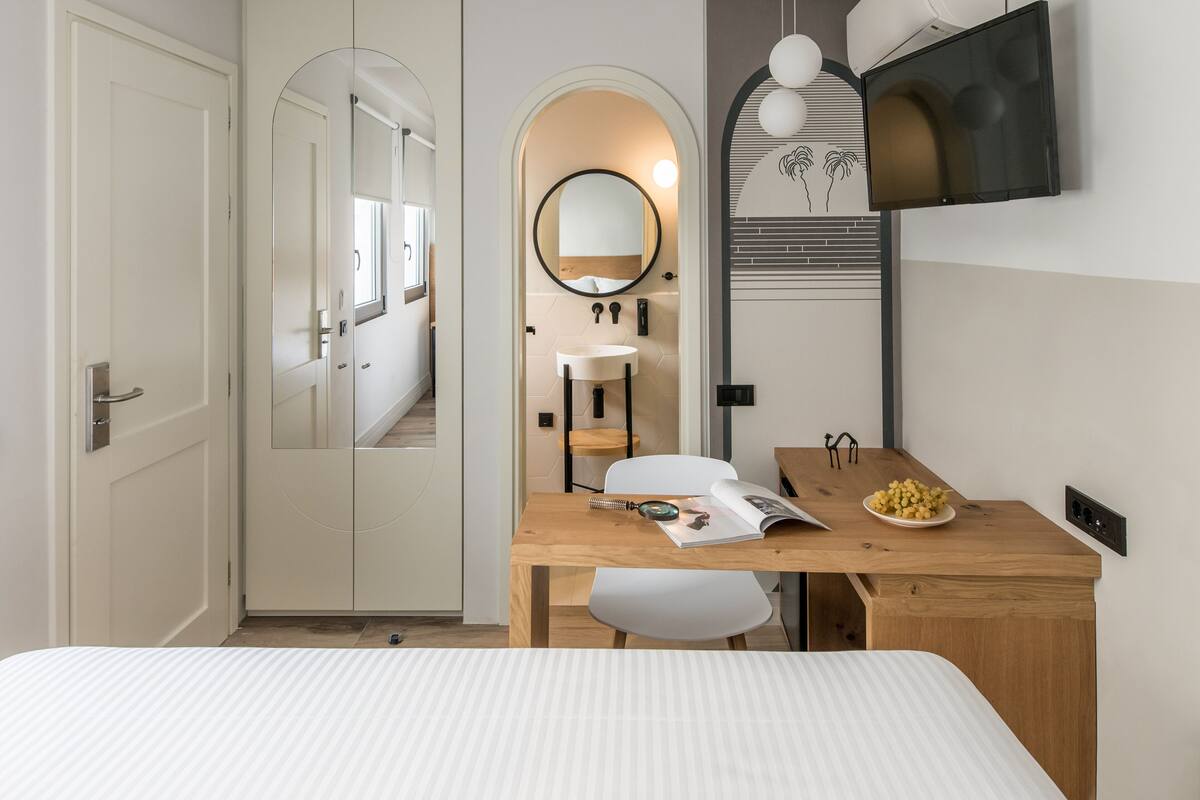 A modern hotel room with a wooden desk, a white chair, a round mirror, and a bed with a striped bedspread.