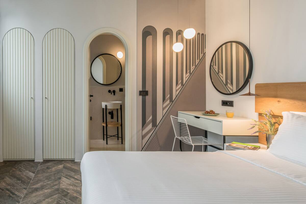 A modern hotel room with a bed, a desk with a chair, a mirror, and a staircase.
