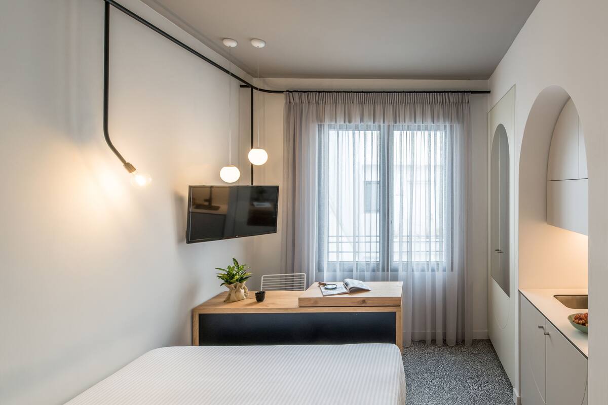 A modern hotel room with a bed, a desk with a chair, a flat-screen TV, a potted plant, and a large window with curtains.