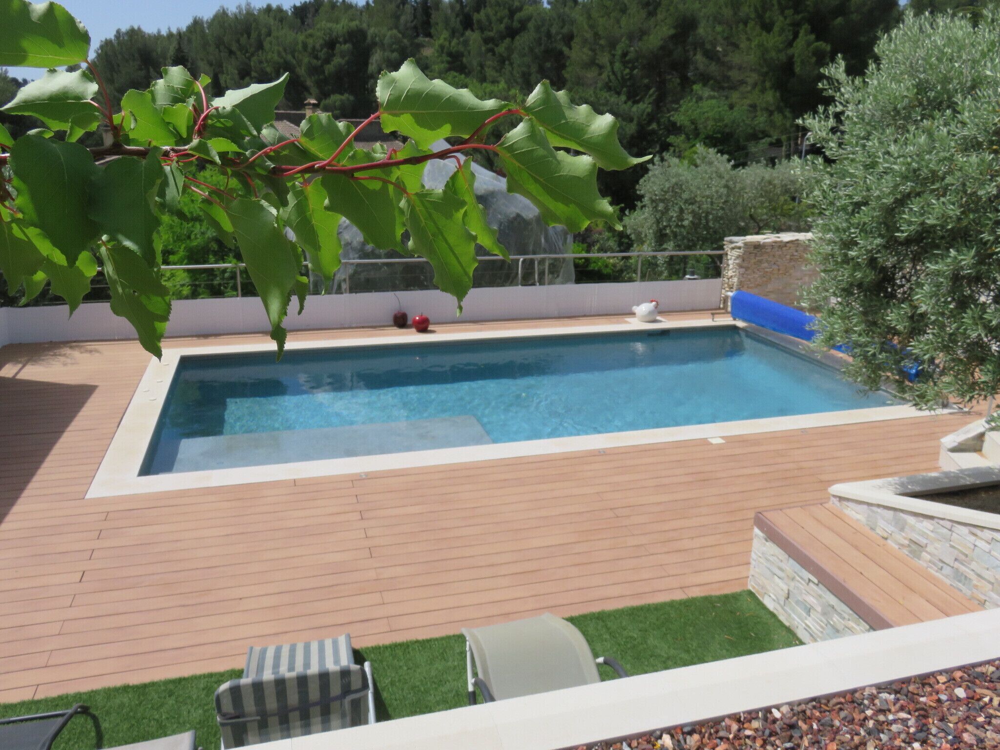 A rectangular swimming pool with a wooden deck, surrounded by greenery and a stone wall.