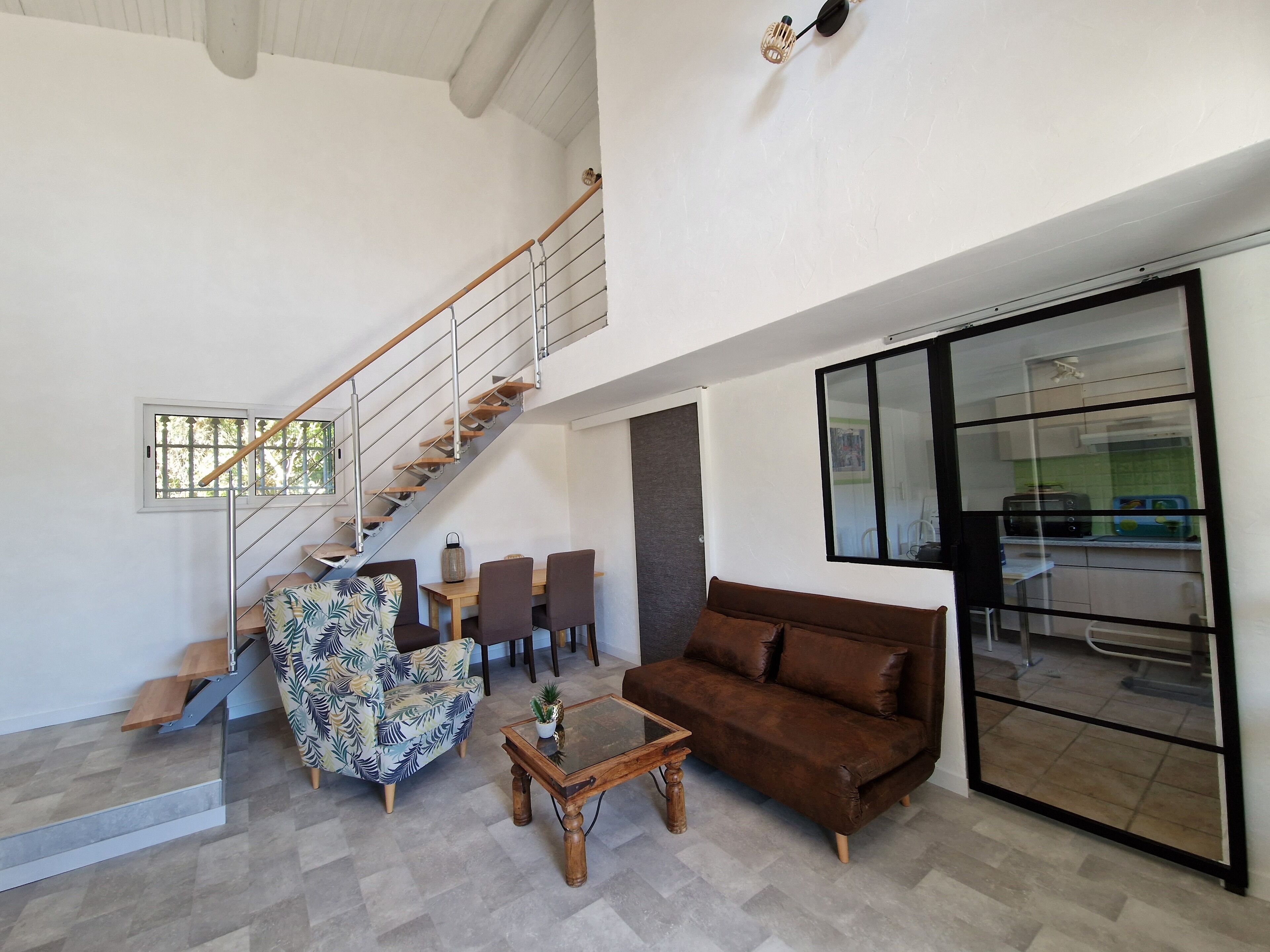 A spacious living area with a staircase, a brown sofa, a patterned armchair, a coffee table, and a view into the kitchen through a glass door.