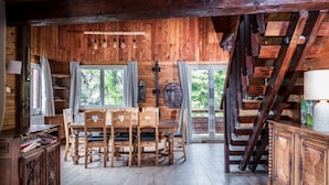 Een rustiek houten blokhutinterieur met een eettafel en stoelen, een trap en uitzicht op de natuur.