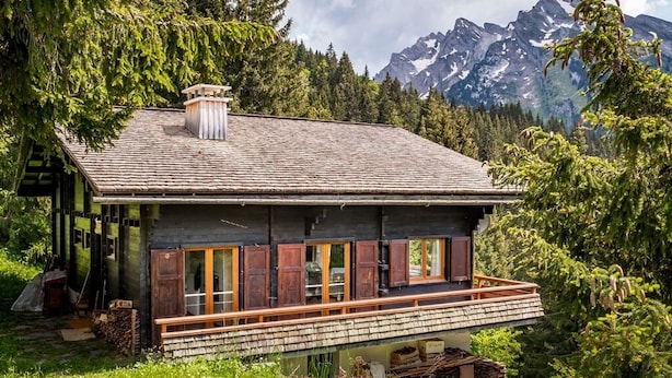 Een rustieke chalet met een houten terras, omringd door weelderige begroeiing en bergen op de achtergrond.