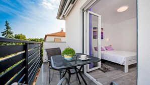 Un balcon avec une table, des chaises, un pot de fleurs et un lit agrémenté d’un coussin violet.