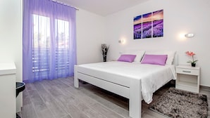 Une chambre moderne avec un grand lit, une table de chevet, une lampe fixée au mur et une vue sur un champ violet à travers des rideaux transparents.