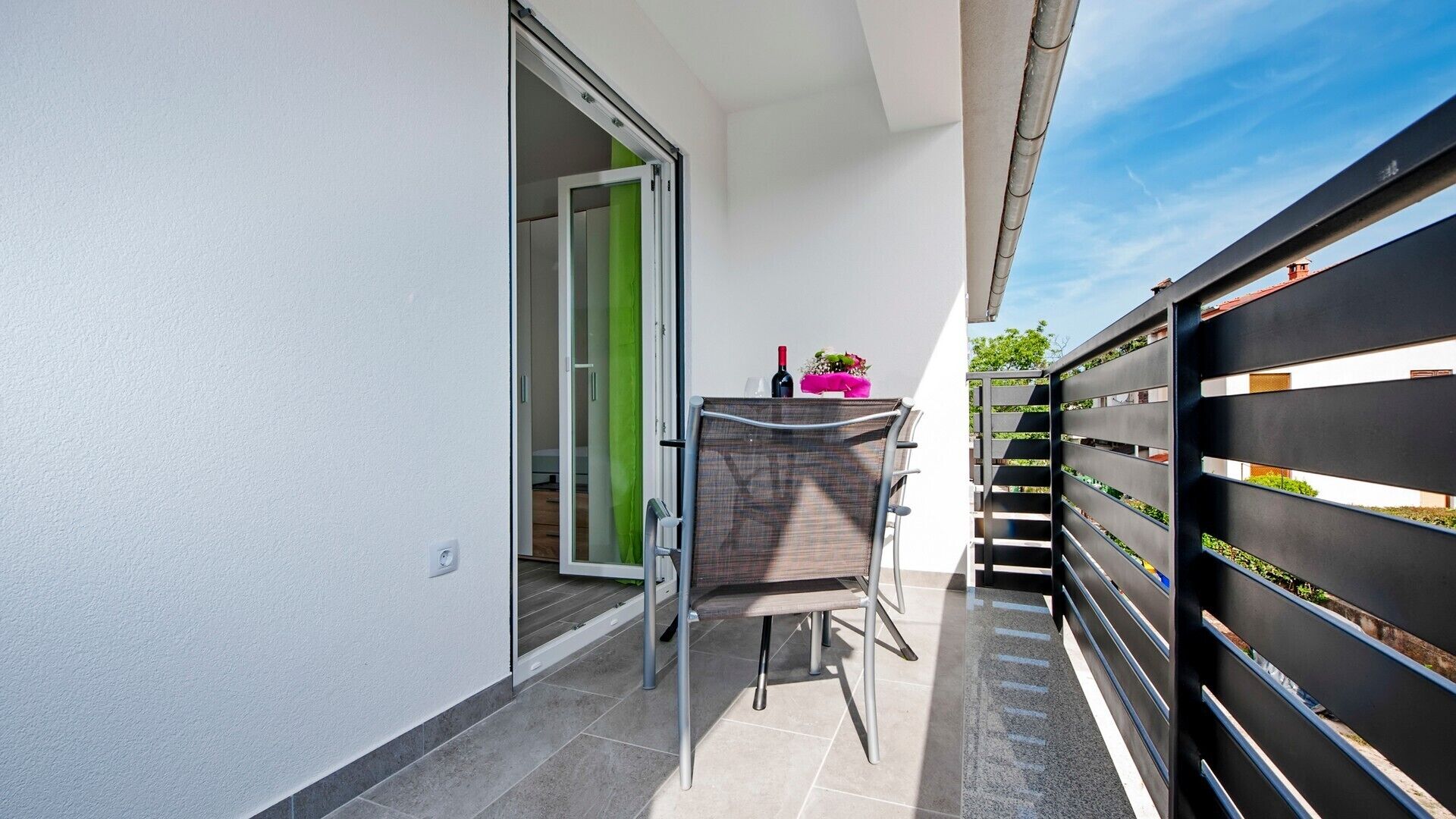 A balcony with a chair, a table with a bottle and flowers, and a black fence with greenery.