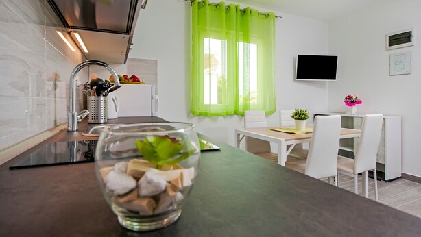 A modern kitchen with a stainless steel range hood, a glass bowl with food items, a dining area with a table and chairs, and a green curtain over the window.