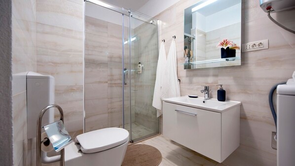 A modern bathroom with a glass shower enclosure, a white toilet, a sink with a soap dispenser, and a mirror above the sink. - ANASTASIA - One Bedroom Apartment (Rovinj)