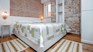 A bedroom with a brick wall, a bed with a quilt, a nightstand, a lamp, and a window. - CASA ROCCIA BIANCA - CIELO - Two Bedroom Apartment (Rovinj)