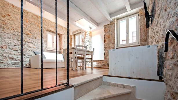 A rustic room with stone walls, a wooden floor, a white sofa, a dining table with chairs, and a staircase leading to another level. - CASA ROCCIA BIANCA - CIELO - Two Bedroom Apartment (Rovinj)